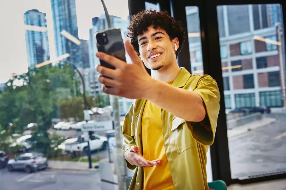 junger Mann blickt lächelnd auf sein Smartphone, welches er mit ausgestreckten Arm vor ihn hält