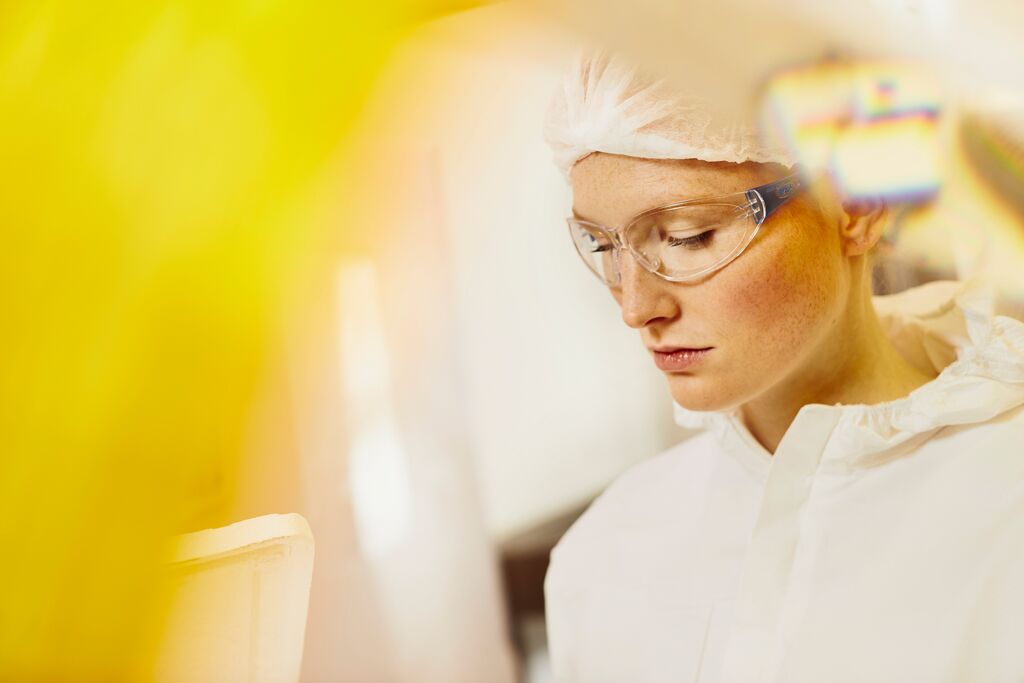 Frau mit weißem Laborkittel, Kopfbedeckung und Brille, die konzentriert nach unten blickt.