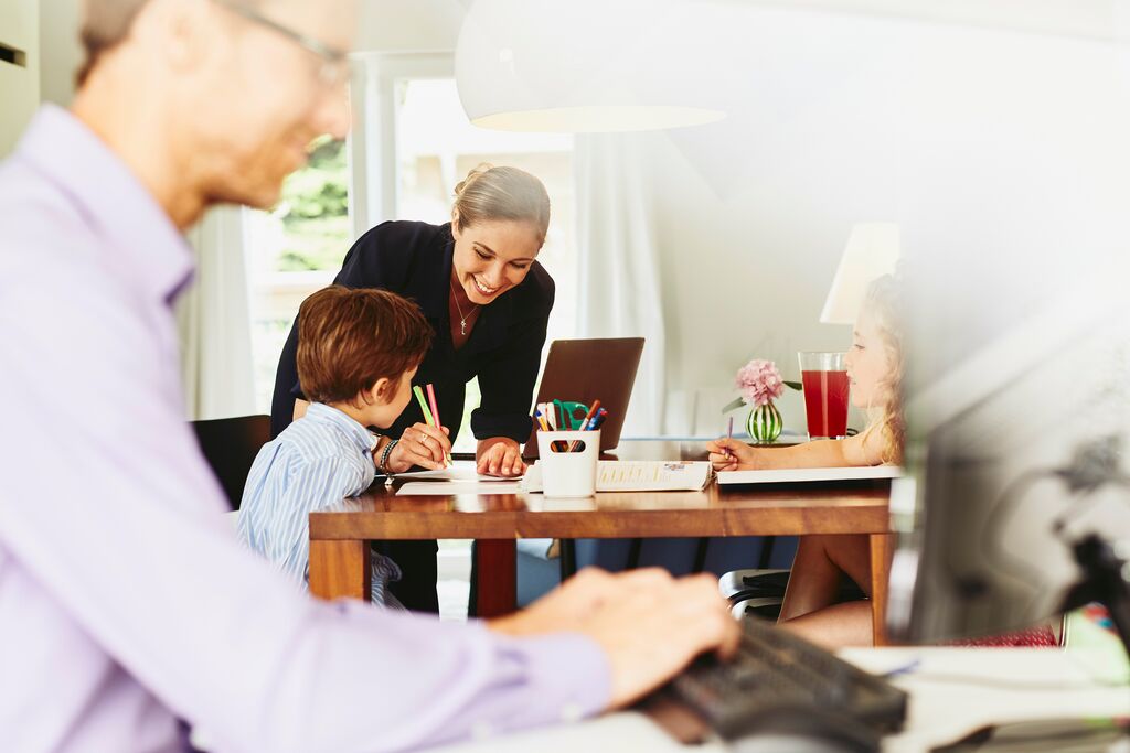 Lächelnde Frau, die ihrem Kind am Küchentisch beim Zeichnen hilft. Im Vordergrund arbeitet ein Mann an einem Computer.