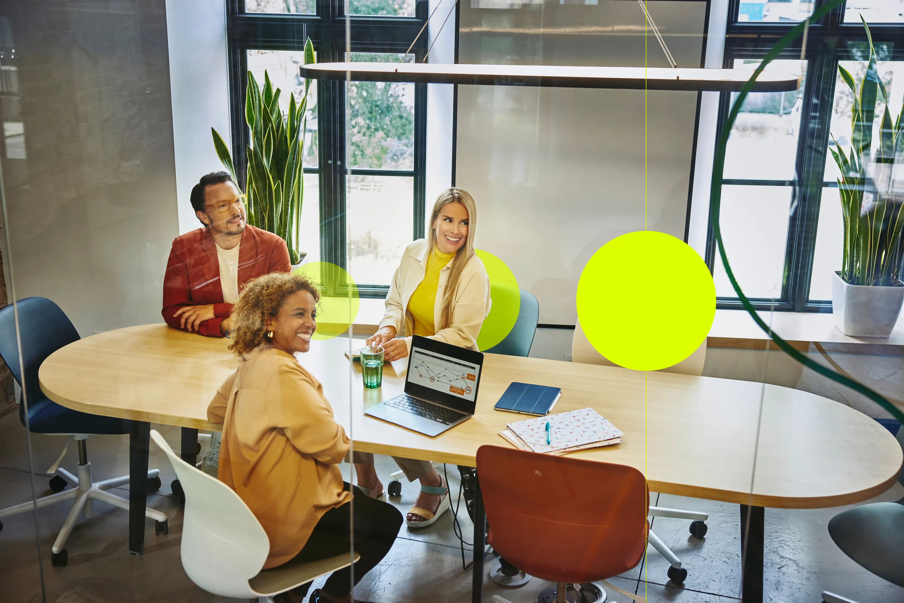 zwei Frauen und ein Mann sitzen an einem Besprechungstisch, blicken in die gleiche Richtung und lächeln