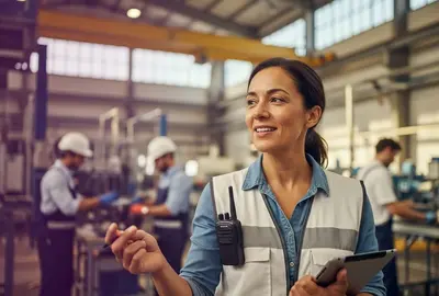eine Frau hält in einer Fabrikhalle ein Tablet in der Hand und ist umgeben von Industrieanlagen