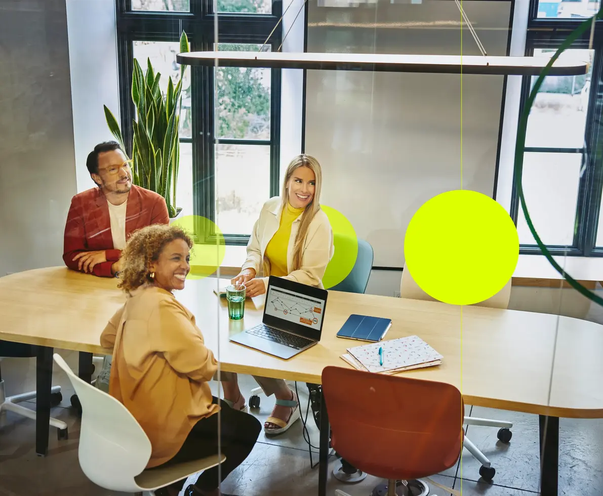 zwei Frauen und ein Mann sitzen an einem Besprechungstisch, blicken in die gleiche Richtung und lächeln