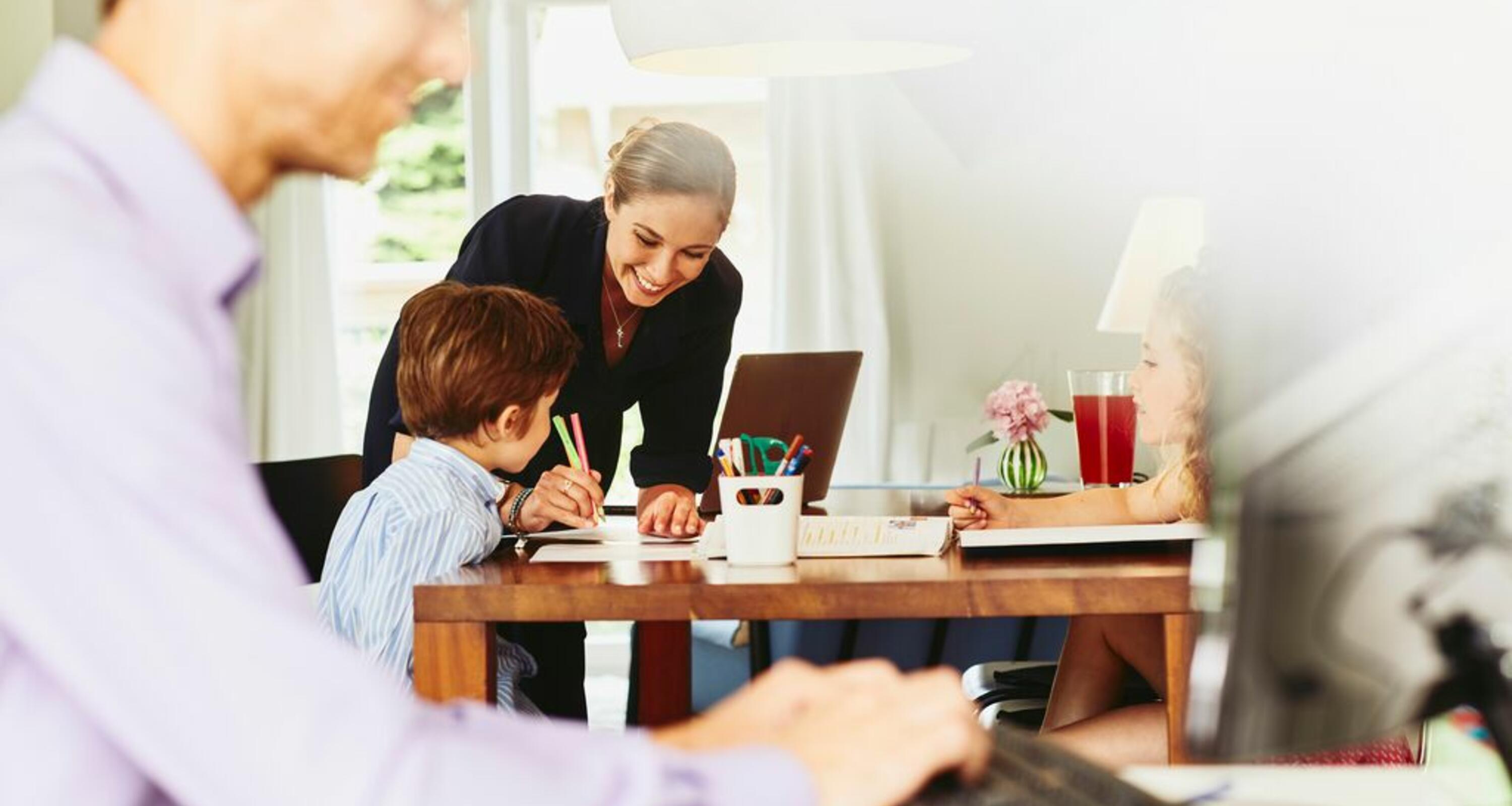 Lächelnde Frau, die ihrem Kind am Küchentisch beim Zeichnen hilft. Im Vordergrund arbeitet ein Mann an einem Computer.