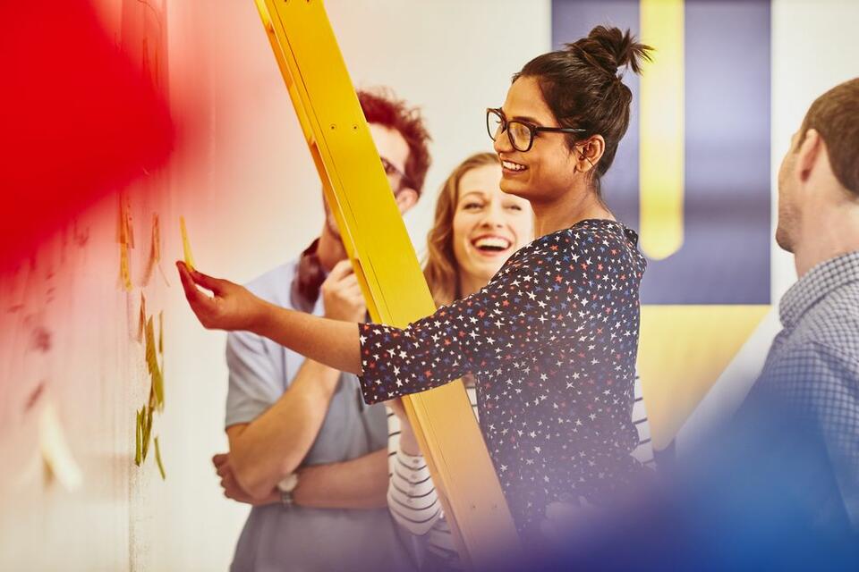 Frau klebt ein Post-it an ein Whiteboard. Um sie herum stehen lächelnde Frauen.