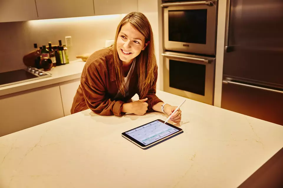 Frau mit Touchpen und Tablet steht in einer Küche und blickt lächelnd zur Seite