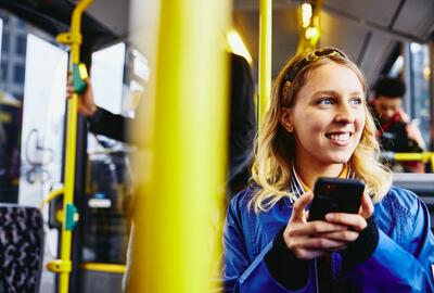Lächelnde Frau, die nach draußen schaut, während sie ihr Telefon in der Hand hält und im Bus sitzt.