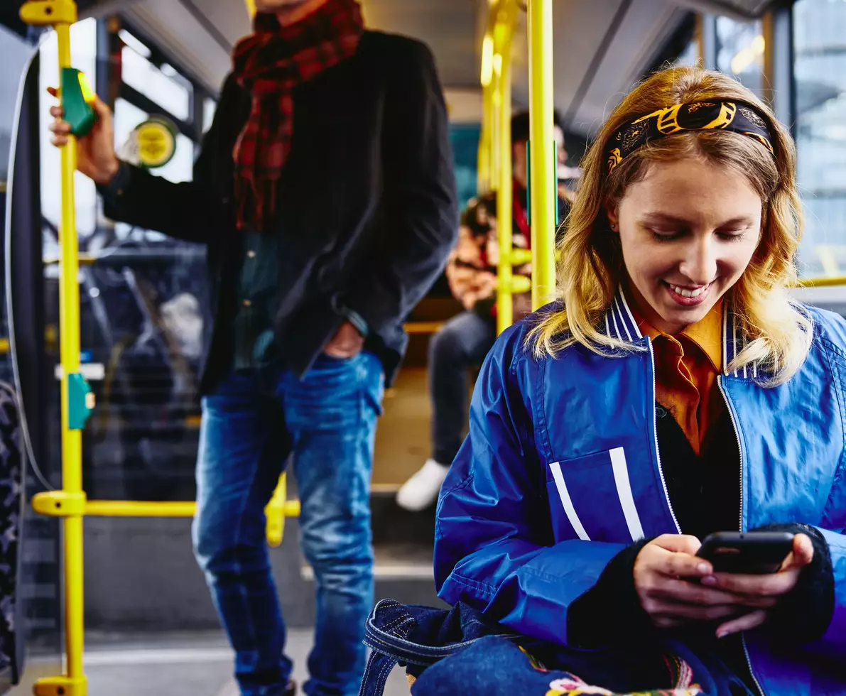 eine junge Frau sitzt in einem Bus und blickt lächelnd auf ihr Smartphone